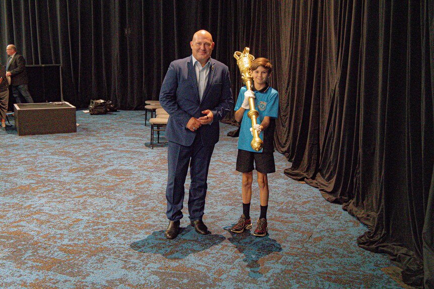 Kid wearing school uniform with mace and man inside a convention centre at a mock-up parliament sitting.