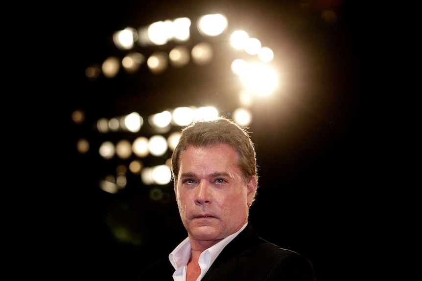 A middle aged man wearing an open-collared white shirt and black jacket appears before some lighting blurred in the background