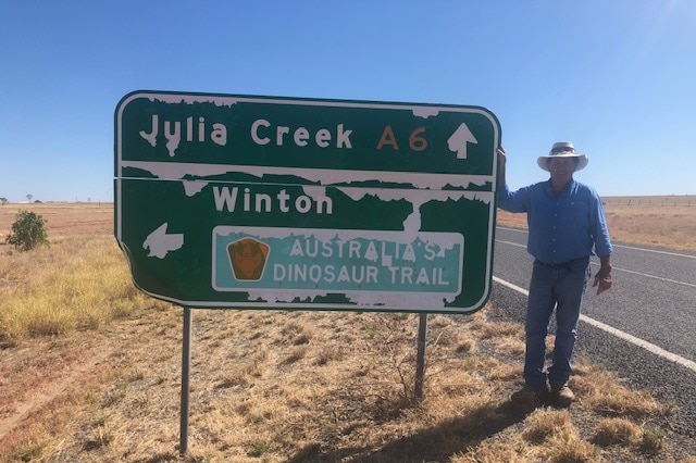 A man wearing jeans and a western-style hat stands next to an aged highway sign. The ground is dry and vast.