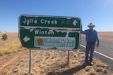 A man wearing jeans and a western-style hat stands next to an aged highway sign. The ground is dry and vast.