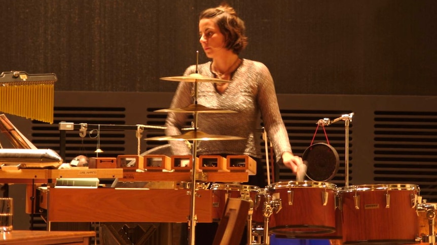 Percussionist in long-sleeved gold top behind bank of percussion (L-R: wind chimes, woodblocks, tom-toms, hanging gong)