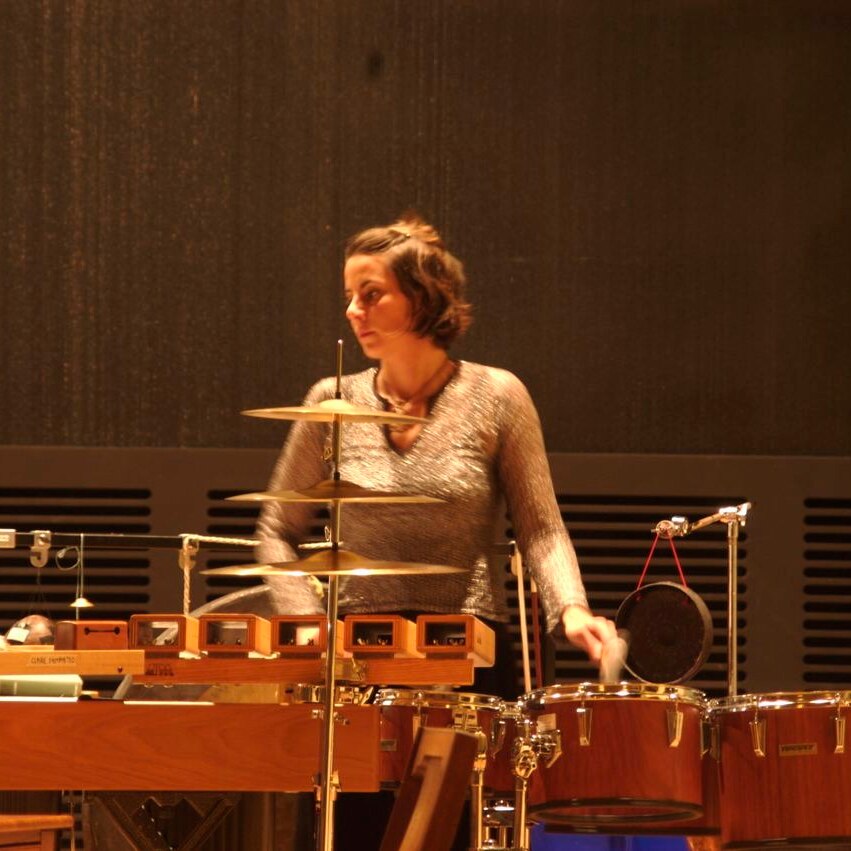 Percussionist in long-sleeved gold top behind bank of percussion (L-R: wind chimes, woodblocks, tom-toms, hanging gong)