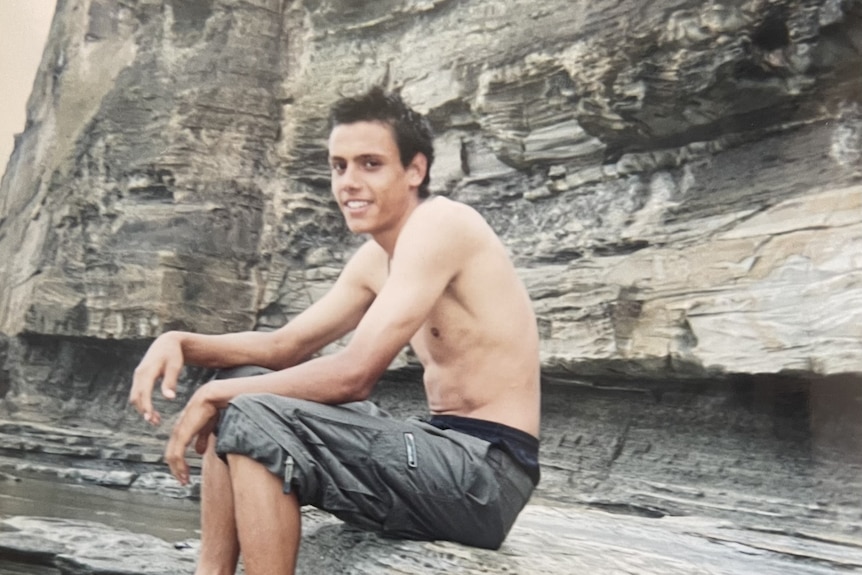 A young Indigenous boy sits shirtless on a rock by the beach.