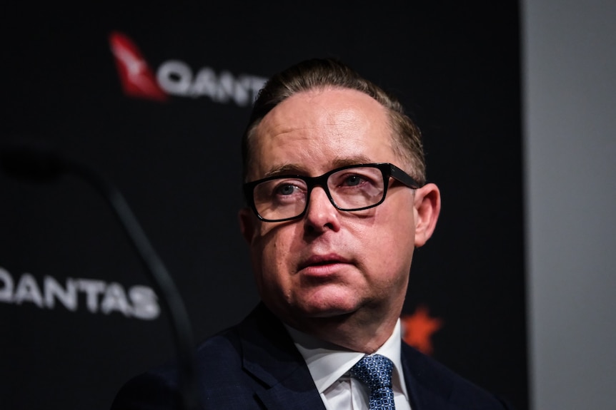 An older man with dark hair and thick rimmed square glasses waring a suit and tie looks ahead in front of Qantas logos.