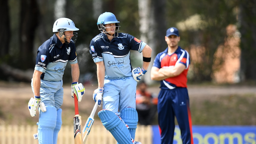 Two men in batting gear stand chatting with a third man wearing red stands with his arms folded in the background