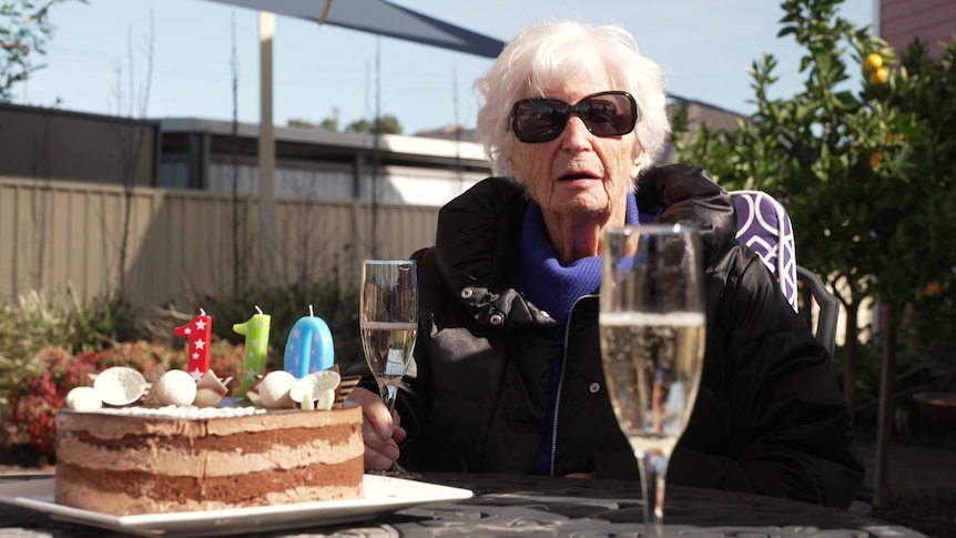 A woman wearing sunglasses sits at a table with a cake and glass of champagne