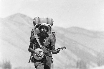 Black & white photo of John Francis walking, carrying a backpack and a banjo