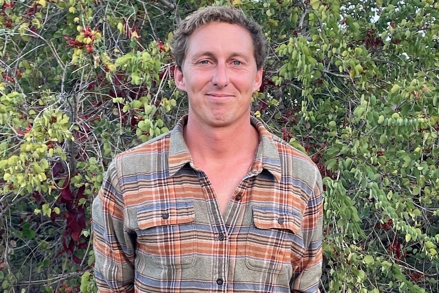 Man with short hair and checkered shirt stands in front of a hedge. 