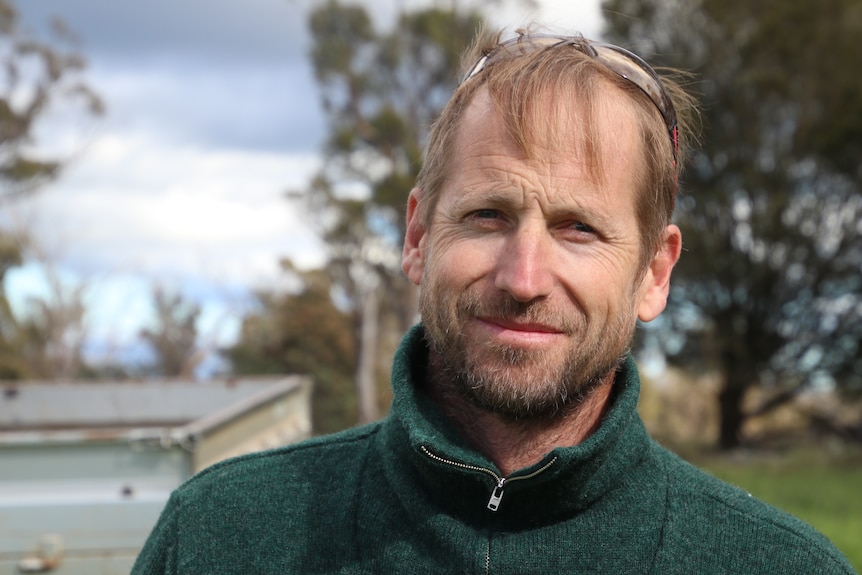 A man in a green jumper with blonde hair and beard stands close to the camera.   