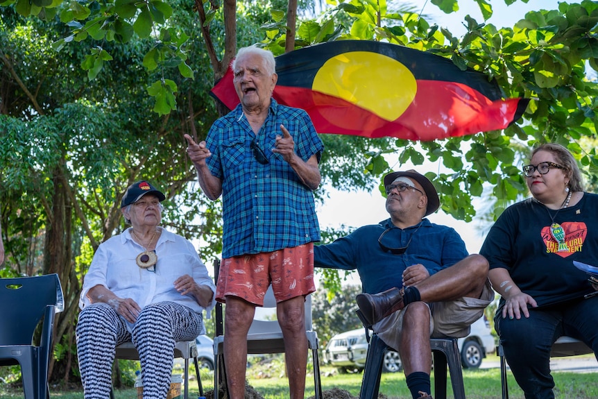 97-year-old "Pop" standing and speaking passionately in front of the Aboriginal flag, during an outdoor meeting.