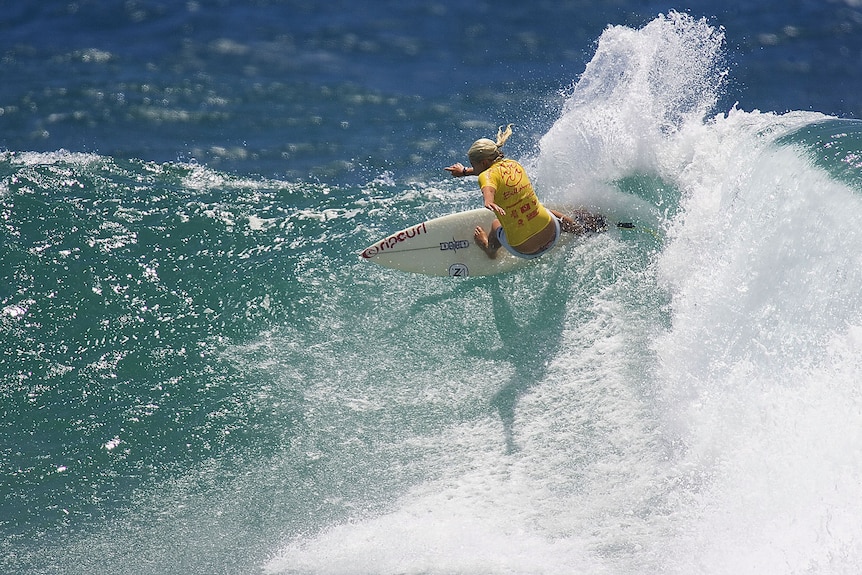 Australian surfer Steph Gilmore turns her board at the top of a wave as white spray flies up during a competition.