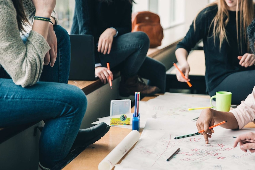 Four women sit around a brainstorming sheet with markers in their hands indicating an enjoyable and collaborative workplace.