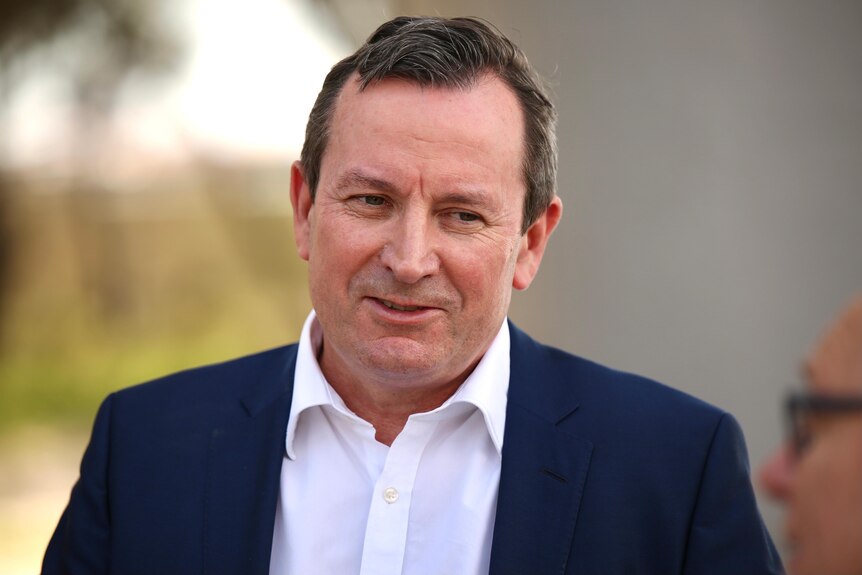 Mark McGowan smiling, outdoors, wearing a blue suit, white shirt, and no tie