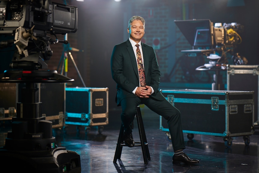 A man wearing a suit sits on a stool surrounded by TV camera and lights.