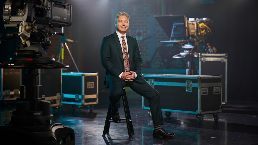 A man wearing a suit sits on a stool surrounded by TV camera and lights.