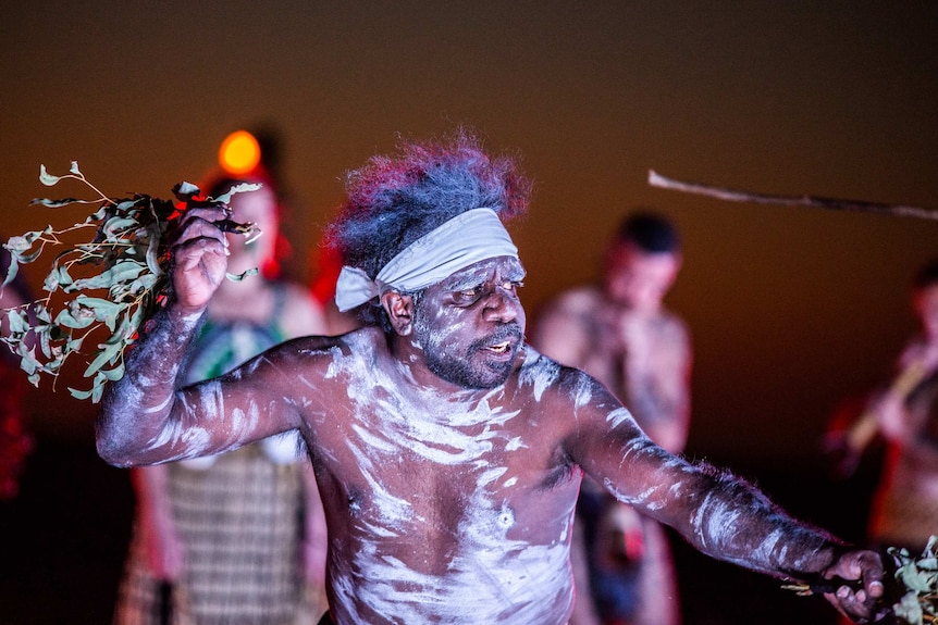 An Indigenous man covered in traditional paint dances.