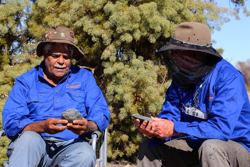 Two men wearing bright blue shirts and Akubra-style hats hold rocks.