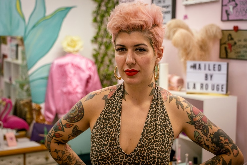 A woman with pink hair and tattoos stands in a pink nail salon.