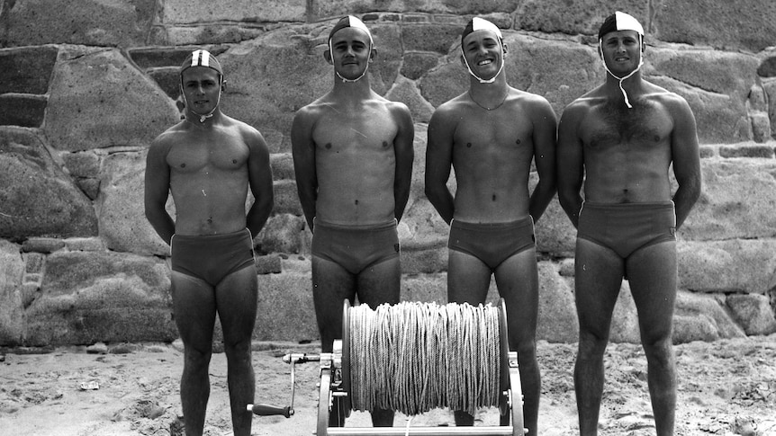 Four lifeguards stand in front of a reel wearing swimming briefs and caps in Jersey.