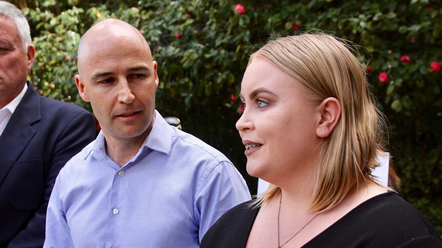 A man in a blue shirt and a woman in a black top hold an interview outside Parliament House.
