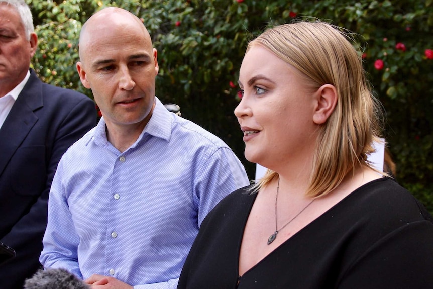 A man in a blue shirt and a woman in a black top hold an interview outside Parliament House.