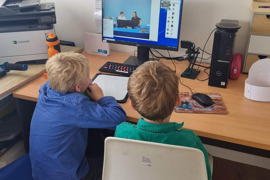 Two boys, one in a blue shirt and one in a green shirt facing a computer screen with two people on it