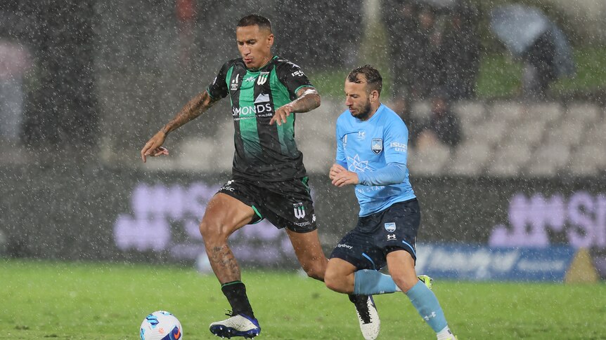 Two male soccer players, one in green and black and another in blue, chase the ball while it rains heavily