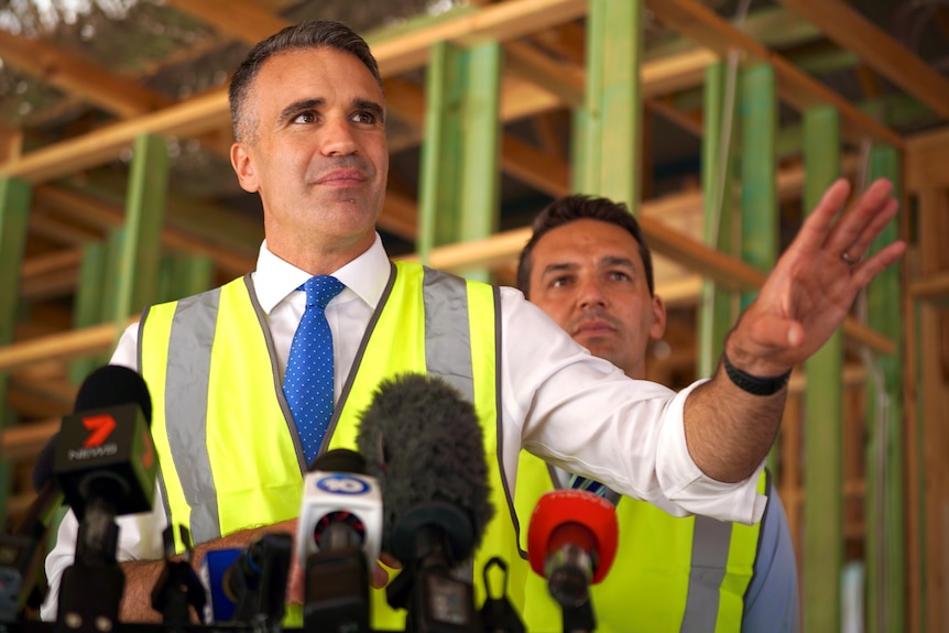 A man wearing a fluoro yellow vest over a business shirt and tie with microphones in front