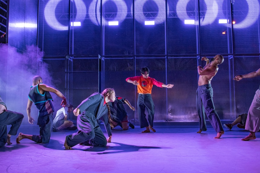 Nine dancers move across a smoky stage, some moving from a crouched position, others on tiptoe