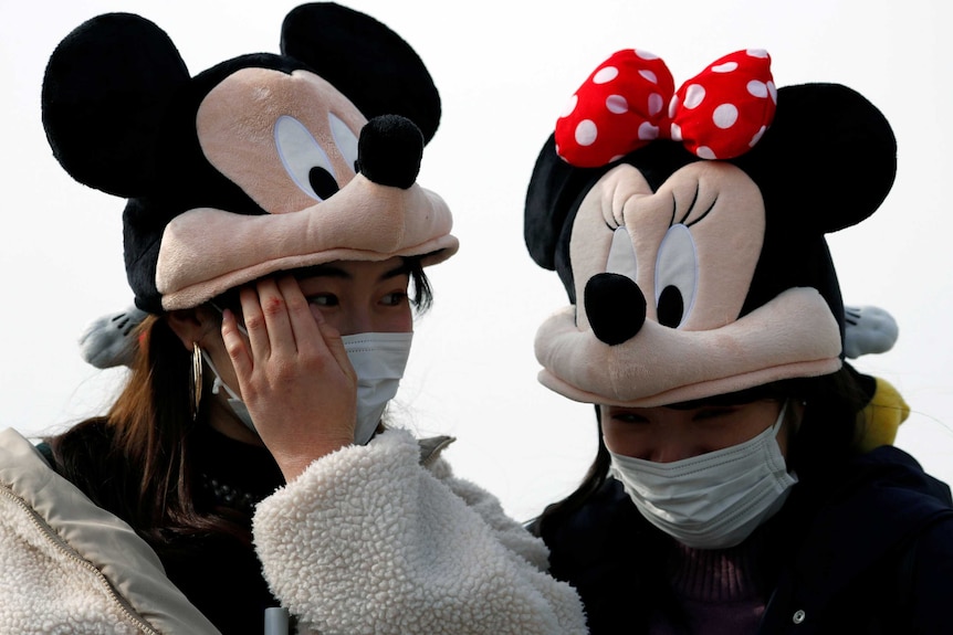 Two people wearing medical masks over their mouths and noses while wearing Micky and Minnie Mouse hats on their heads.