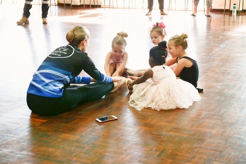 A young woman and four little girls in ballet costumes stretching on the floor before class.