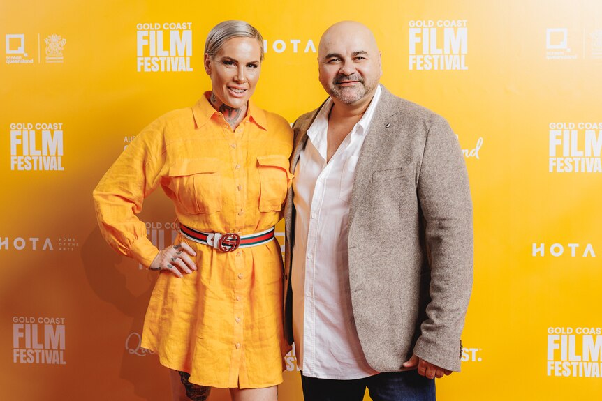 A woman in a yellow dress and a man in a suit stand in front of a wall that reads "Gold Coast Film Festival".