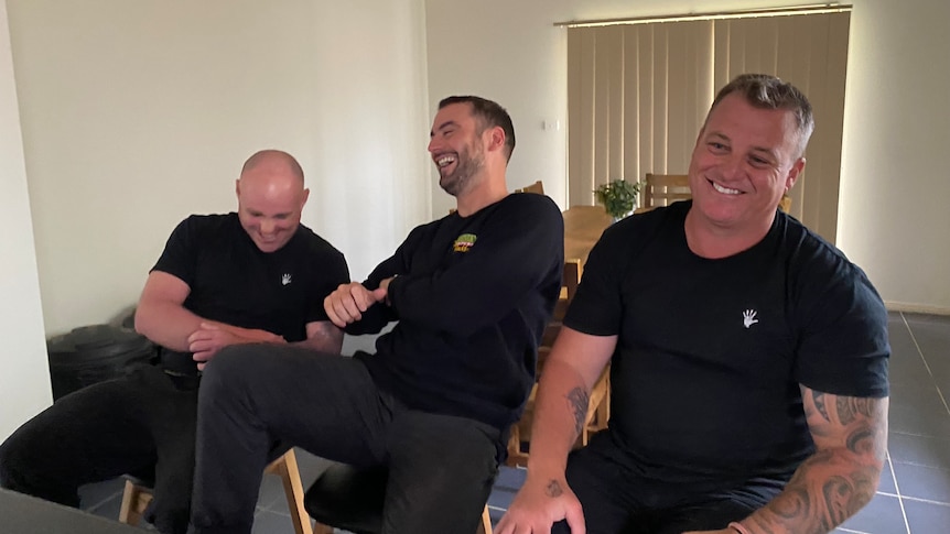 Three men laugh while sitting in a living room, all dressed in black, with logos, one man has tattoos on his arms.