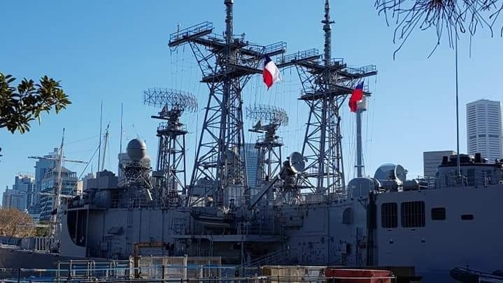 A war ship with Chilean flags on it sits in a harbour on a clear blue sky day.