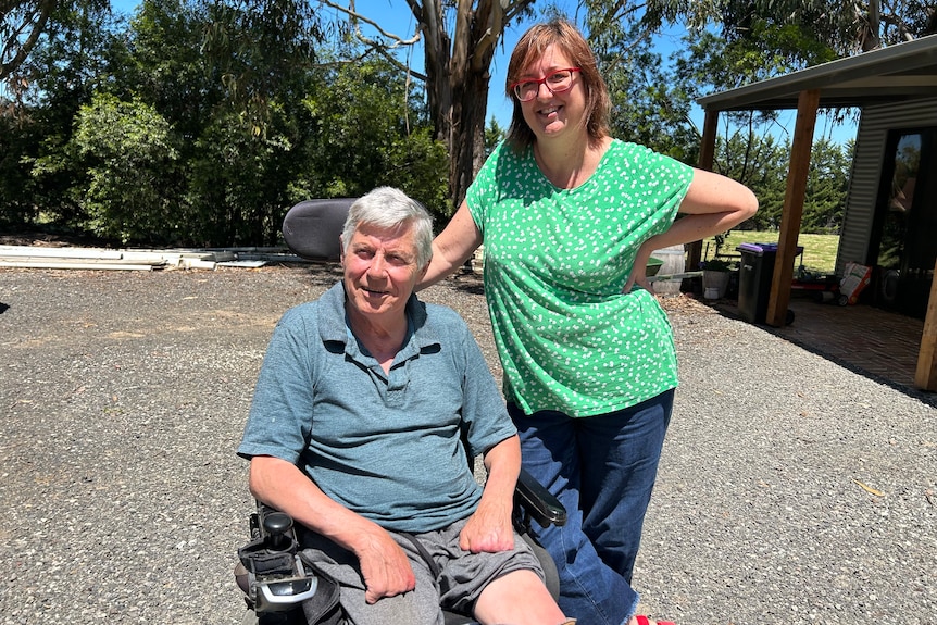 A white man with grey hair sitting in a wheelchair and a woman with brown hair standing up out the front of a farm house