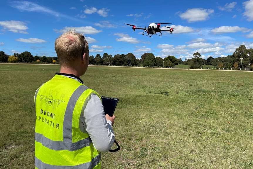 A drone in the background with a man in a yellow coat operating it using a tablet.