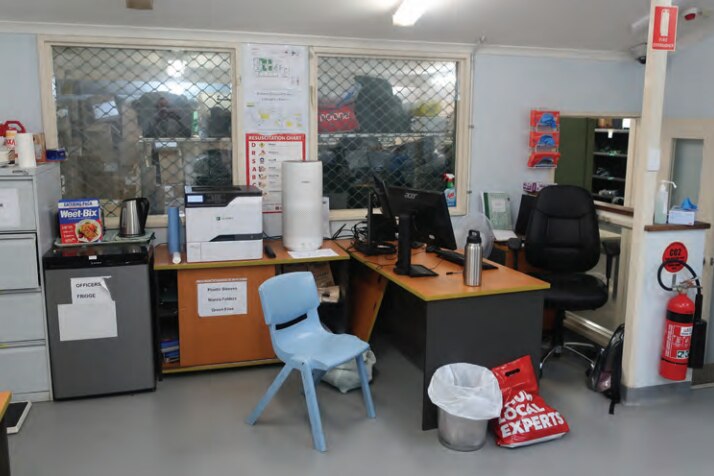 An small, old, slightly cluttered looking office with a desk, computer, plastic chairs, fridge and filing cabinets.