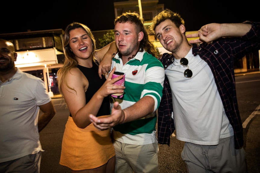 Two young men and a woman embrace each other in a platonic way on a sidewalk during a night out