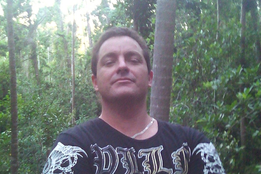 Man stands with arms crossed in a forest looking down at the camera. 