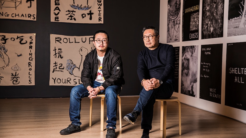 Colour photo of artists Jason Phu and John Young Zerunge sitting in front of their respective artworks for Burrangong Affray.