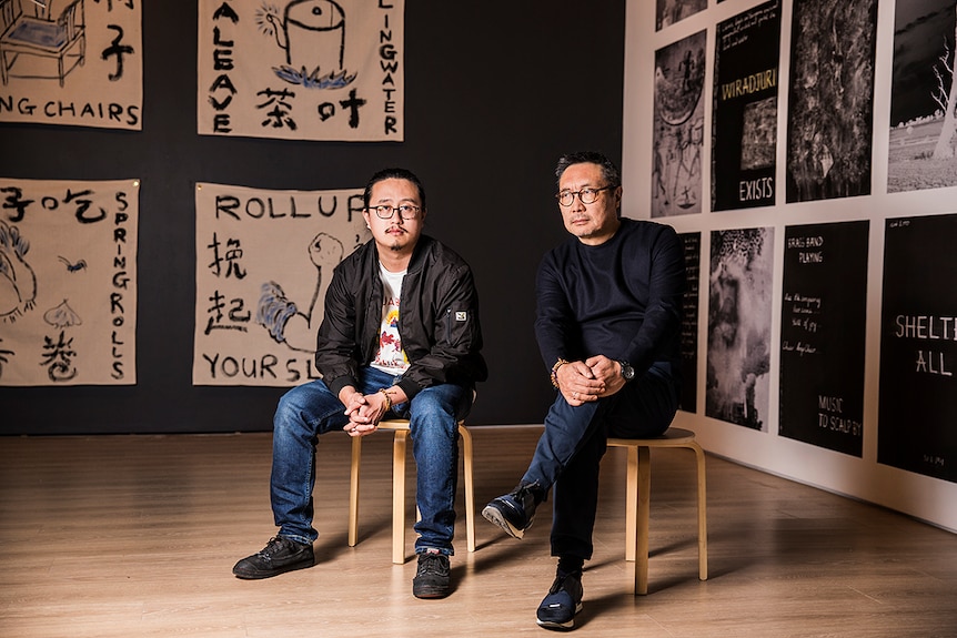 Colour photo of artists Jason Phu and John Young Zerunge sitting in front of their respective artworks for Burrangong Affray.