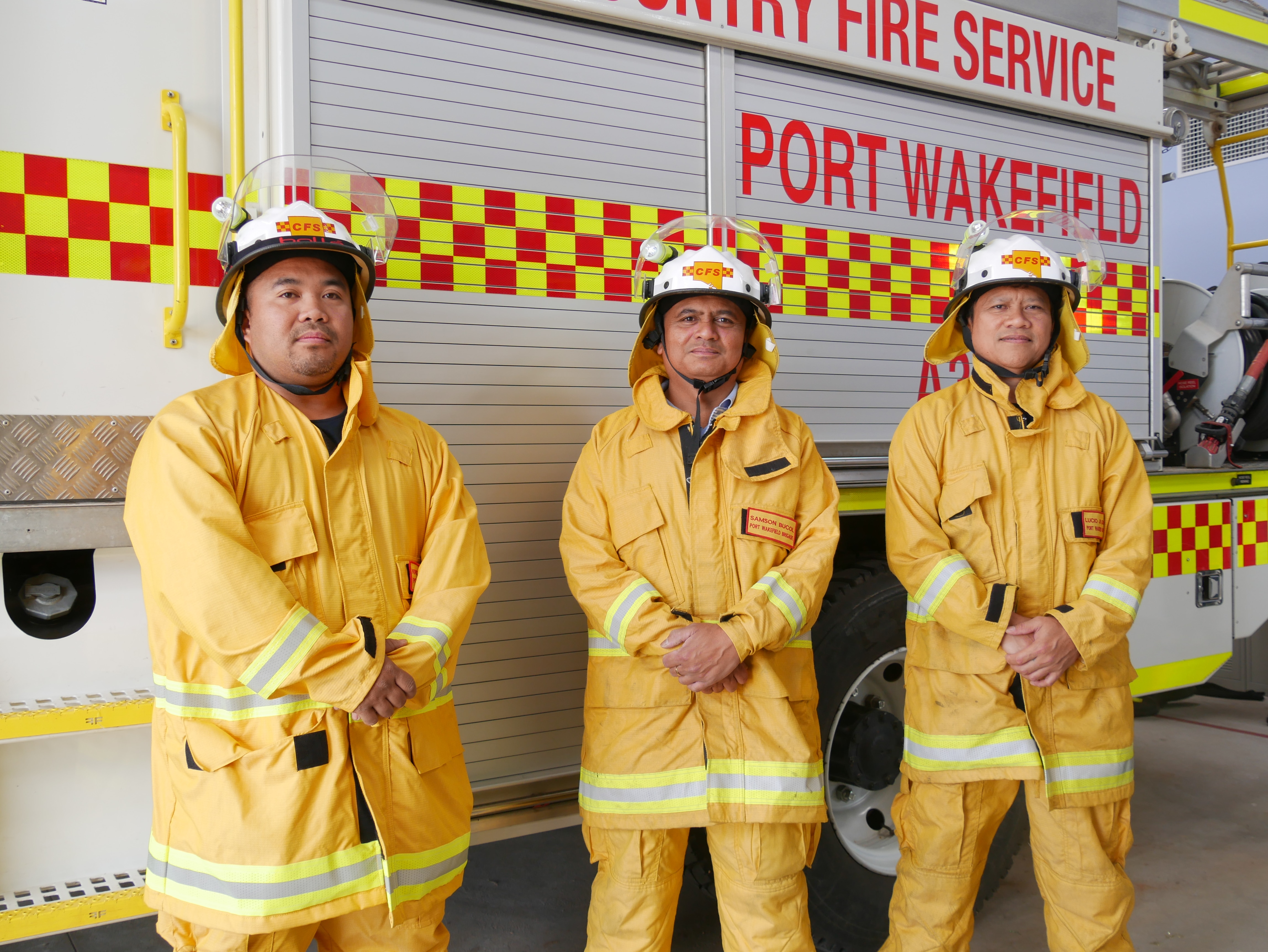 The men in yellow firefighting uniforms and helmets stand in front of a white fire truck. 