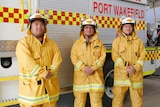 The men in yellow firefighting uniforms and helmets stand in front of a white fire truck. 
