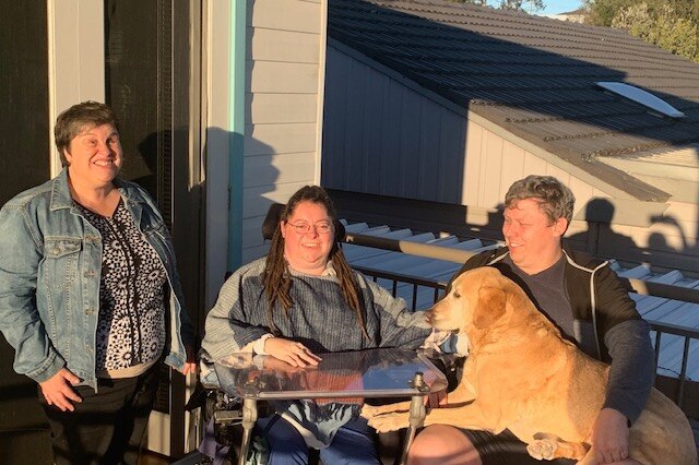 Image of three people and a dog, posing for photographs in the afternoon sun on a suburban deck.