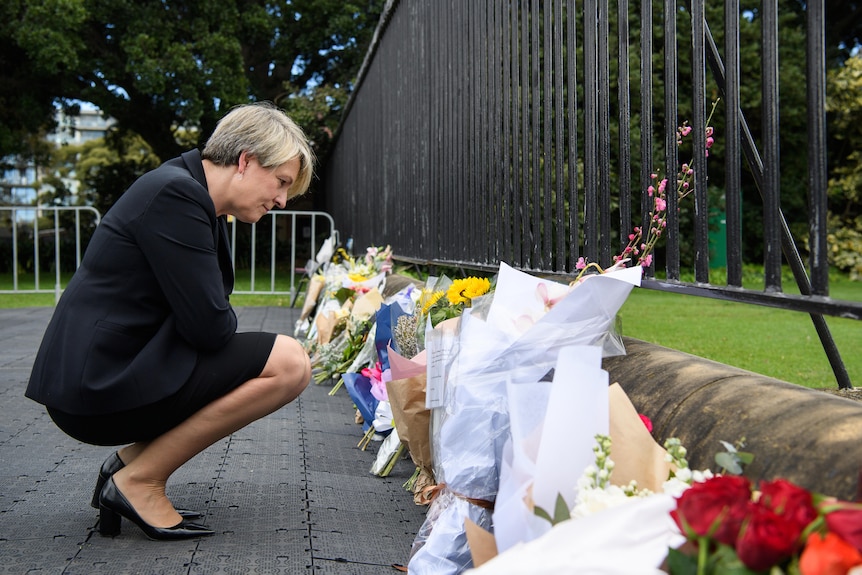 Tanya Plibersek crouches down to look at bouqets of flowers left at the fence of Government House