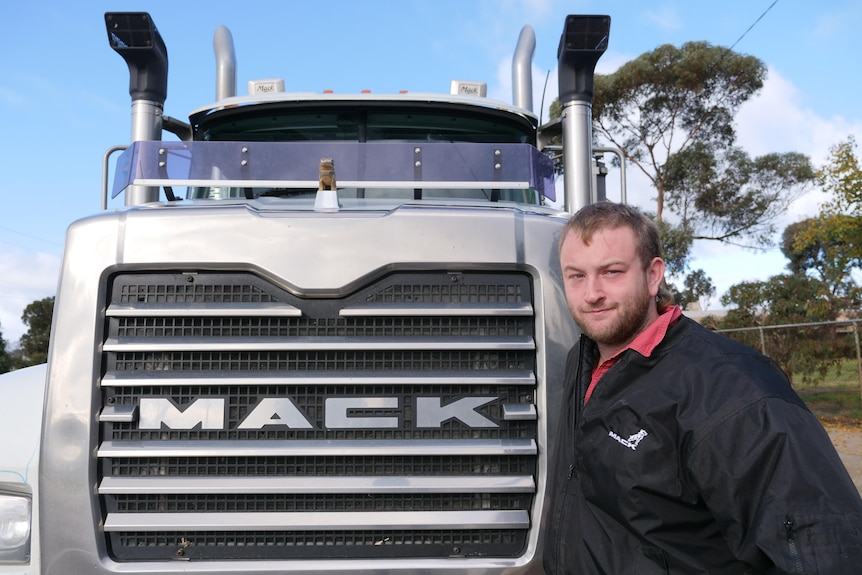 Man with dark blonde hair and bullet with black jacket and red polo stands next to truck