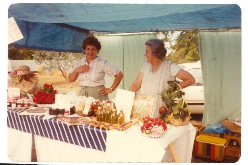 Two women stand behind a table covered with jam jars and pickled vegetables at an outdoor marquee.