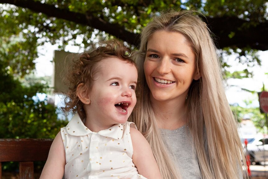 Blonde mother and brunette child smiling outside in front of a tree