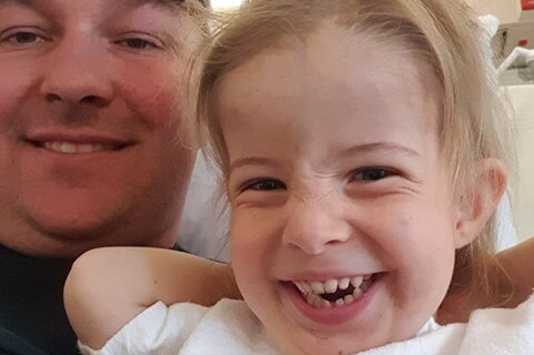 A selfie-style shot of a little blonde girl grinning, with her dad in the background.