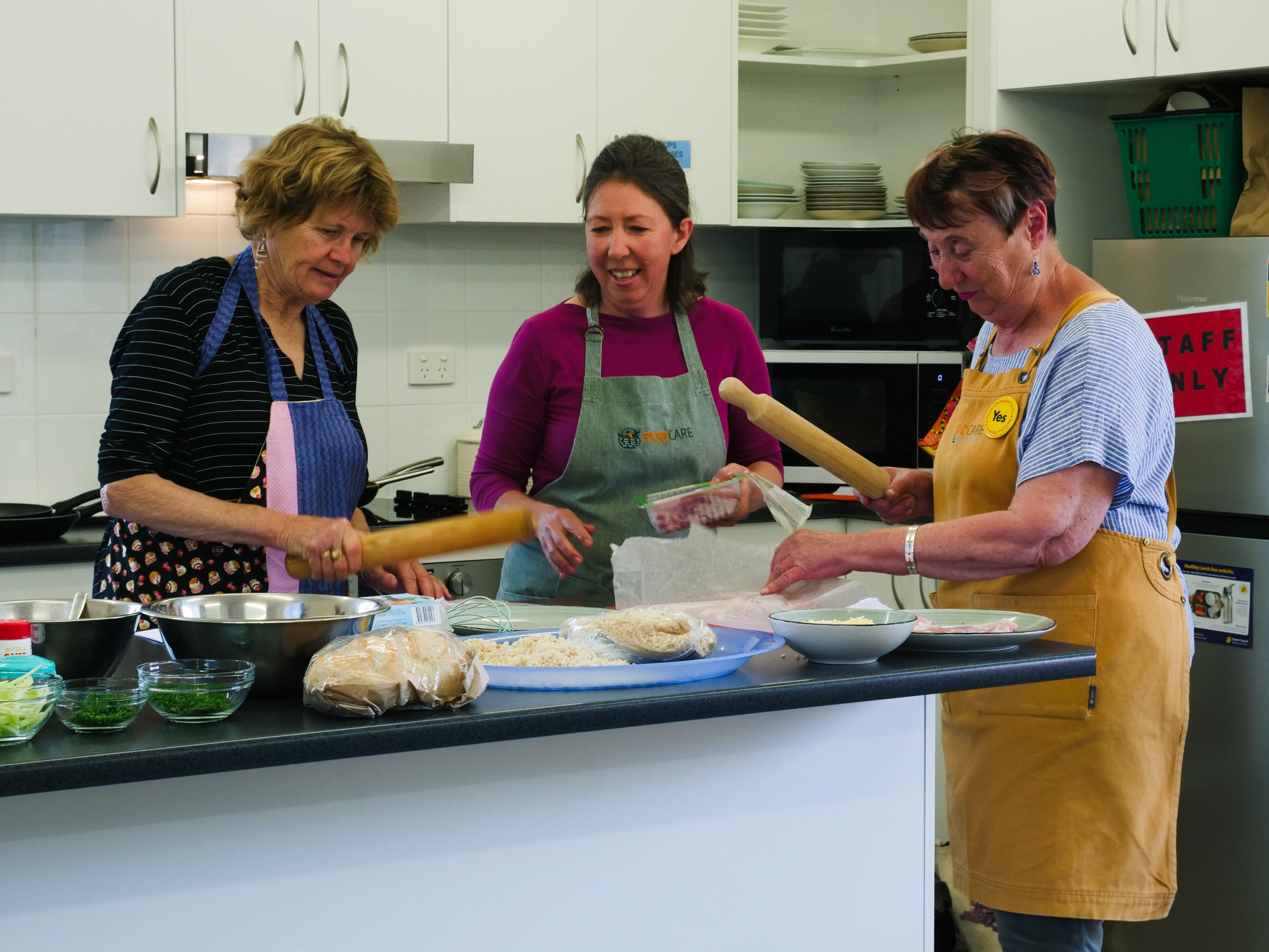Three women in aprons stand behind a kitchen bench. Two are holding rolling pins.